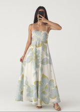 Woman wearing a floral dress taking a mirror selfie against a white curtain.